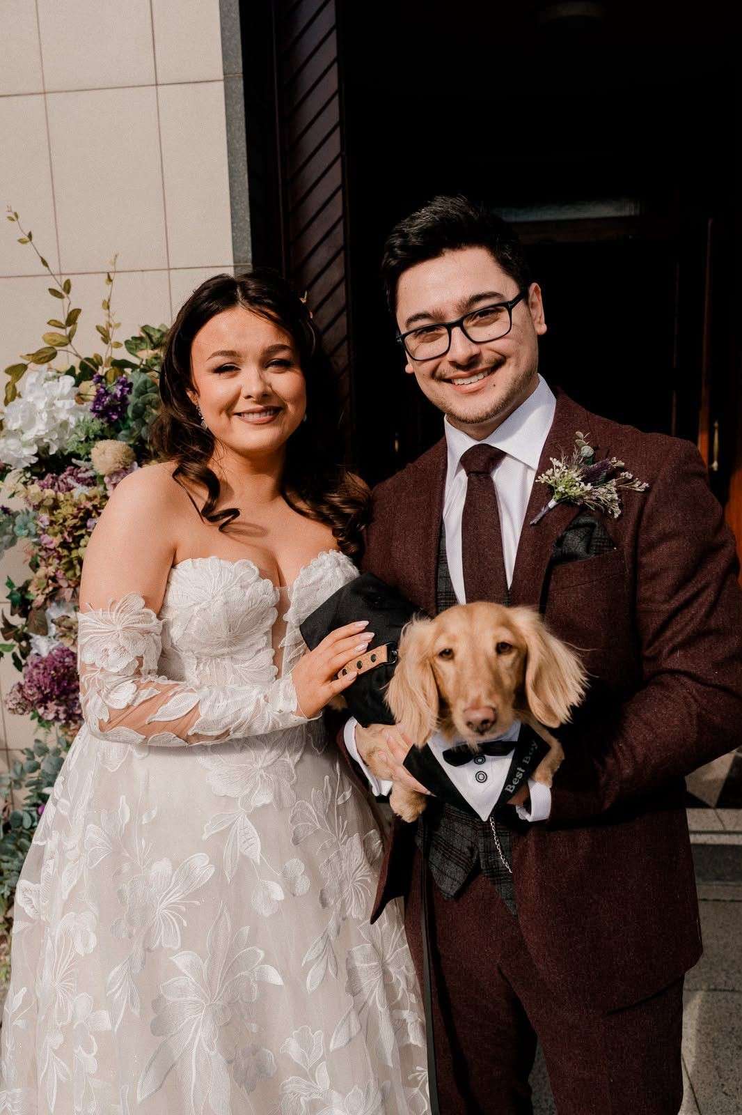 A bride and groom smiling with their dachshund, who wears a black tuxedo bandana with a bow tie